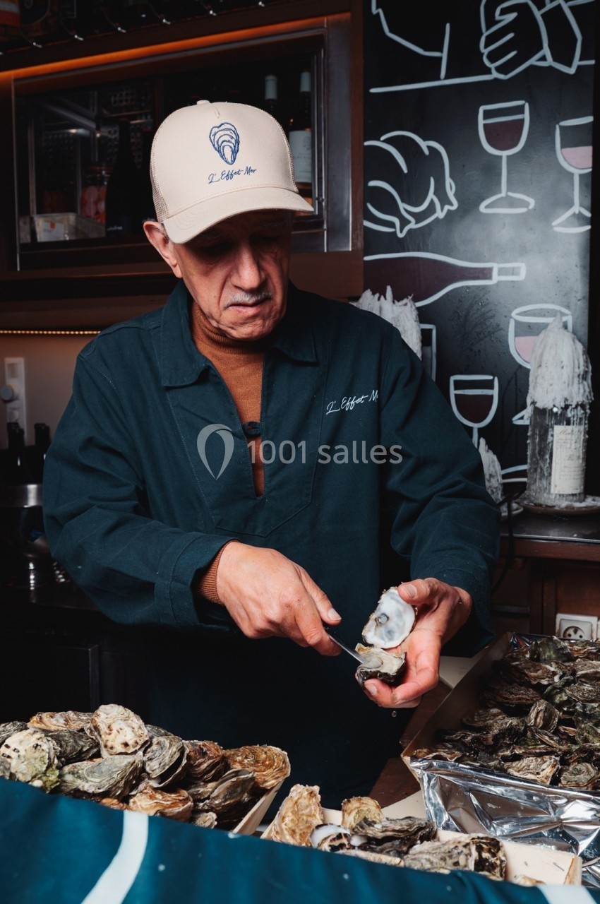 Un homme ouvre une huître dans un étal de fruits de mer, avec des coquilles et un décor de bar en arrière-plan.