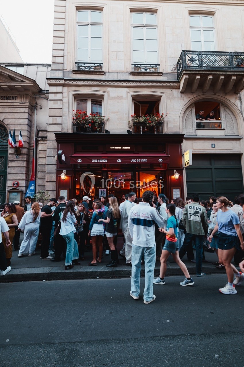 Groupe de personnes rassemblées devant un bar à l'entrée éclairée, sur une rue bordée de bâtiments anciens.