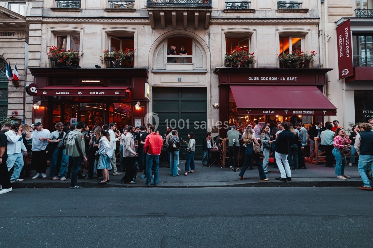 Foule rassemblée devant des bars avec terrasses dans une rue animée, en fin de journée.