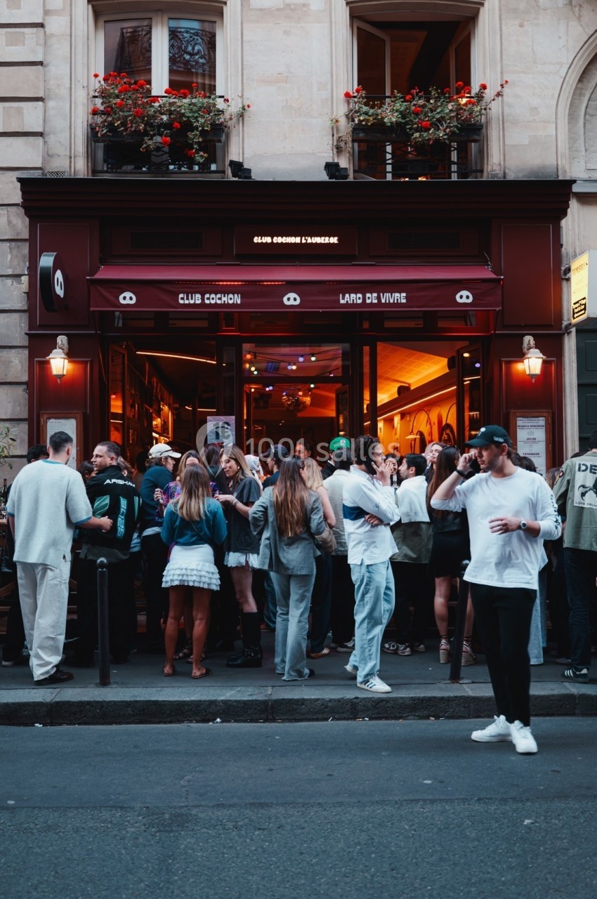 Groupe de personnes rassemblées devant un bar à la façade rouge, avec des fenêtres décorées de fleurs.