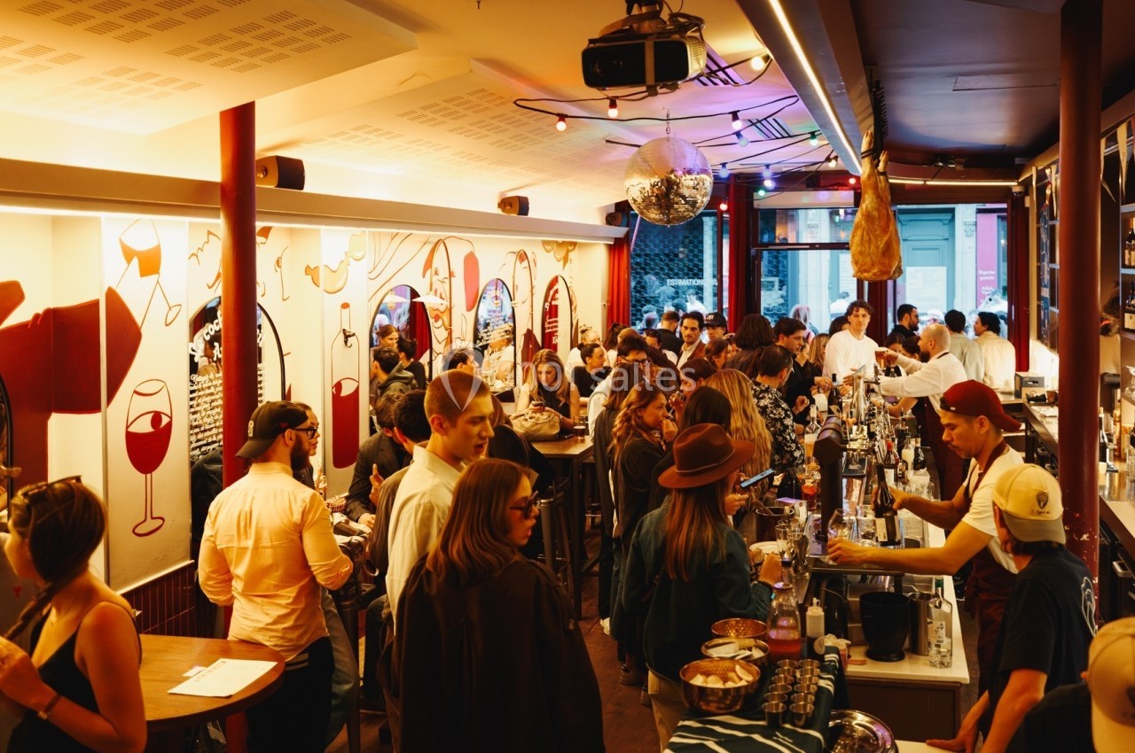 Bar animé avec des clients debout, discutant et commandant des boissons dans une ambiance chaleureuse et éclairée.