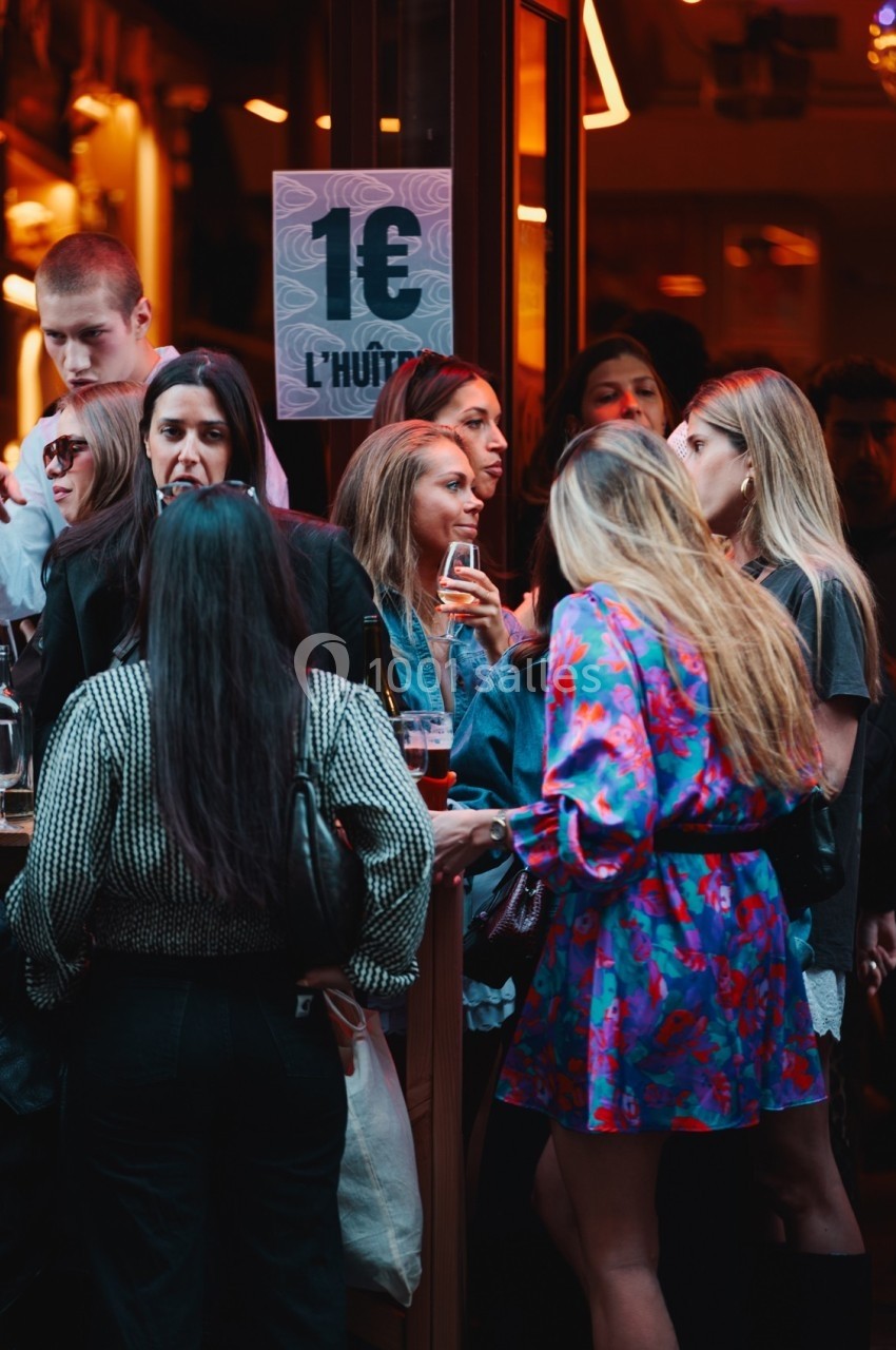 Groupe de personnes discutant et buvant devant un bar animé, avec une affiche indiquant ’1€ l'huître’.