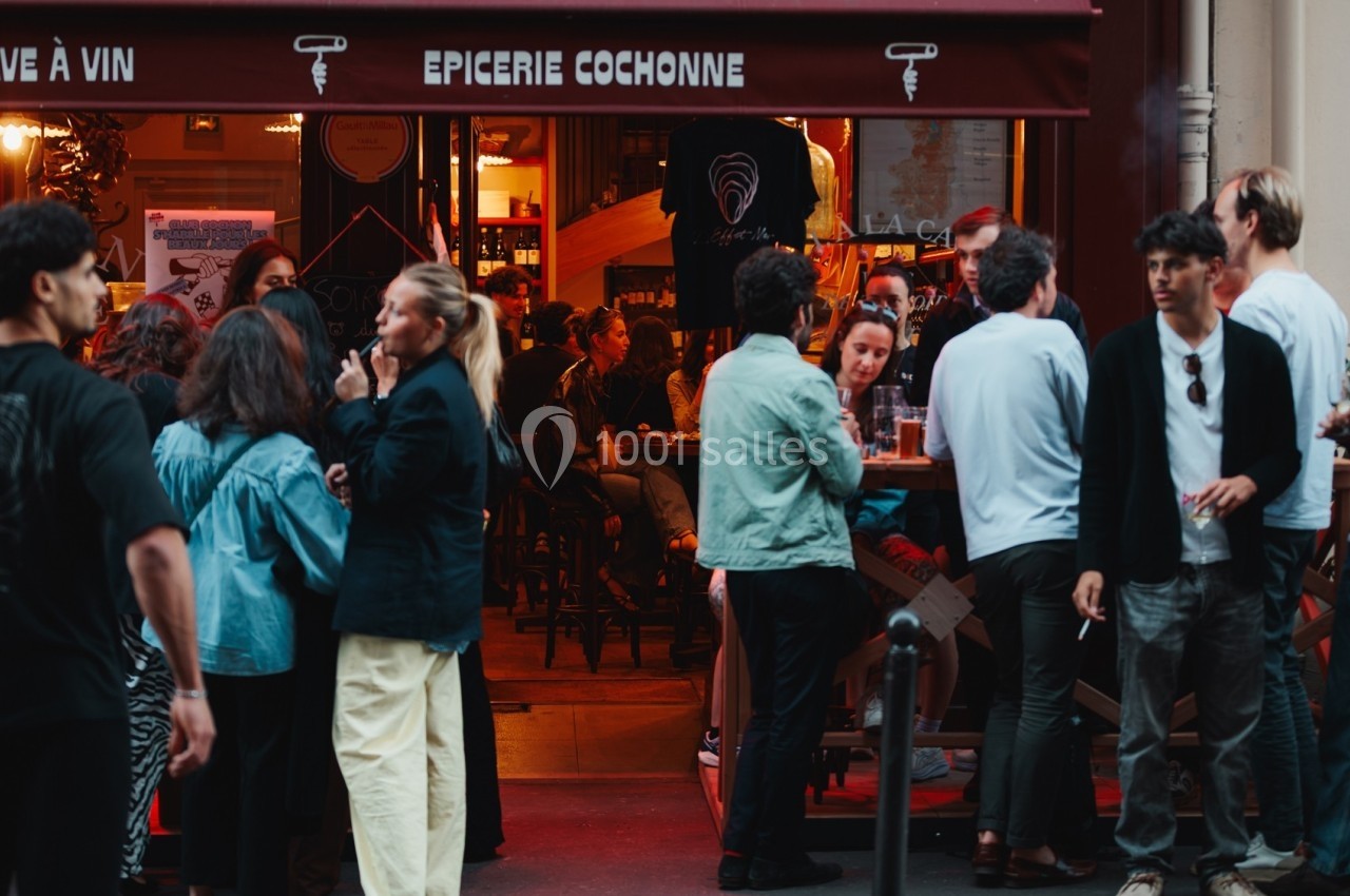 Groupe de personnes rassemblées devant un bar animé avec une enseigne rouge indiquant ’Épicerie Cochonne’.