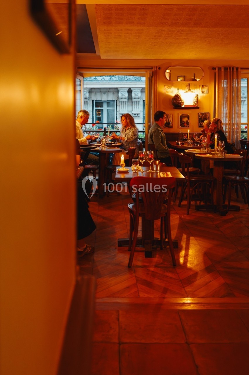 Salle de restaurant chaleureuse avec des clients dînant à des tables en bois, éclairée par une lumière tamisée.