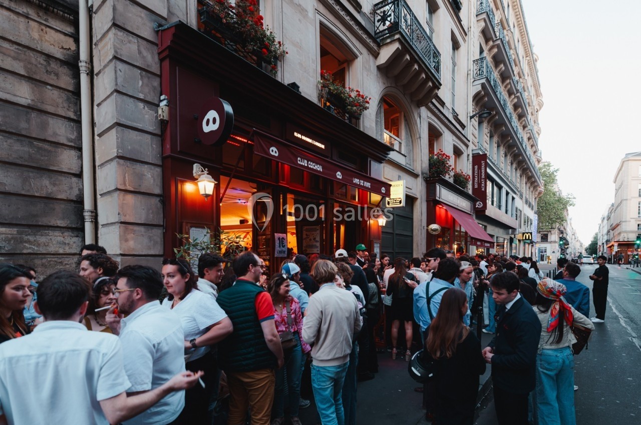 Groupe de personnes rassemblées devant un bar animé dans une rue bordée d'immeubles parisiens.