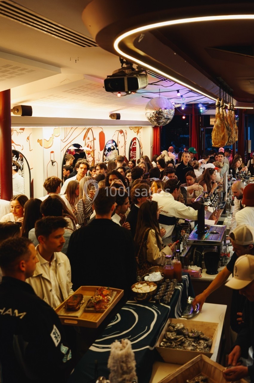 Foule de personnes dans un bar animé, avec un comptoir chargé de plats et boissons, éclairage chaleureux.