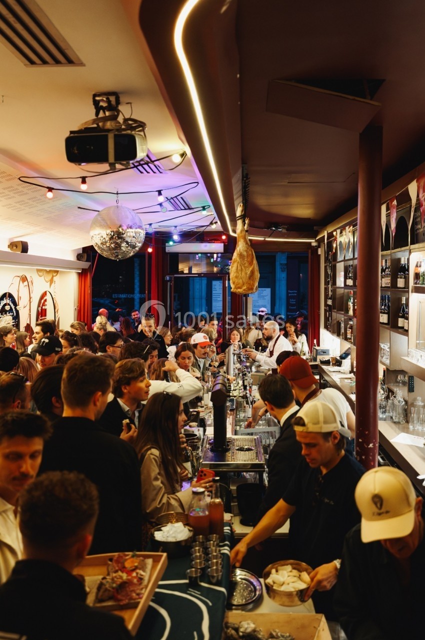 Bar animé en soirée, avec des clients debout discutant et des serveurs préparant des boissons derrière le comptoir.