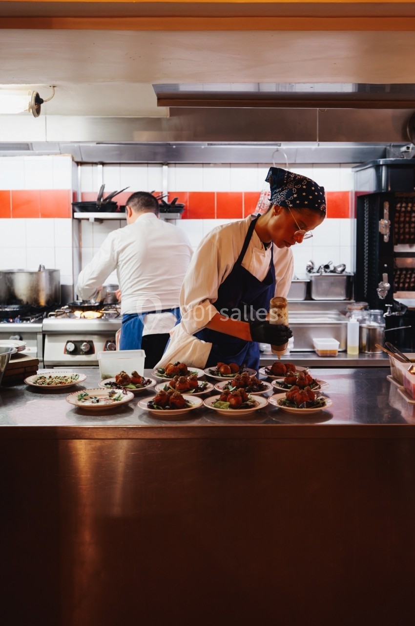 Une cuisinière prépare des assiettes dans une cuisine professionnelle, un autre cuisinier travaille en arrière-plan.