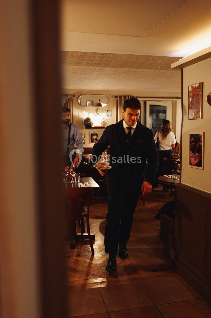 Un serveur en veste sombre marche dans un restaurant chaleureux avec des tables dressées et des clients en arrière-plan.