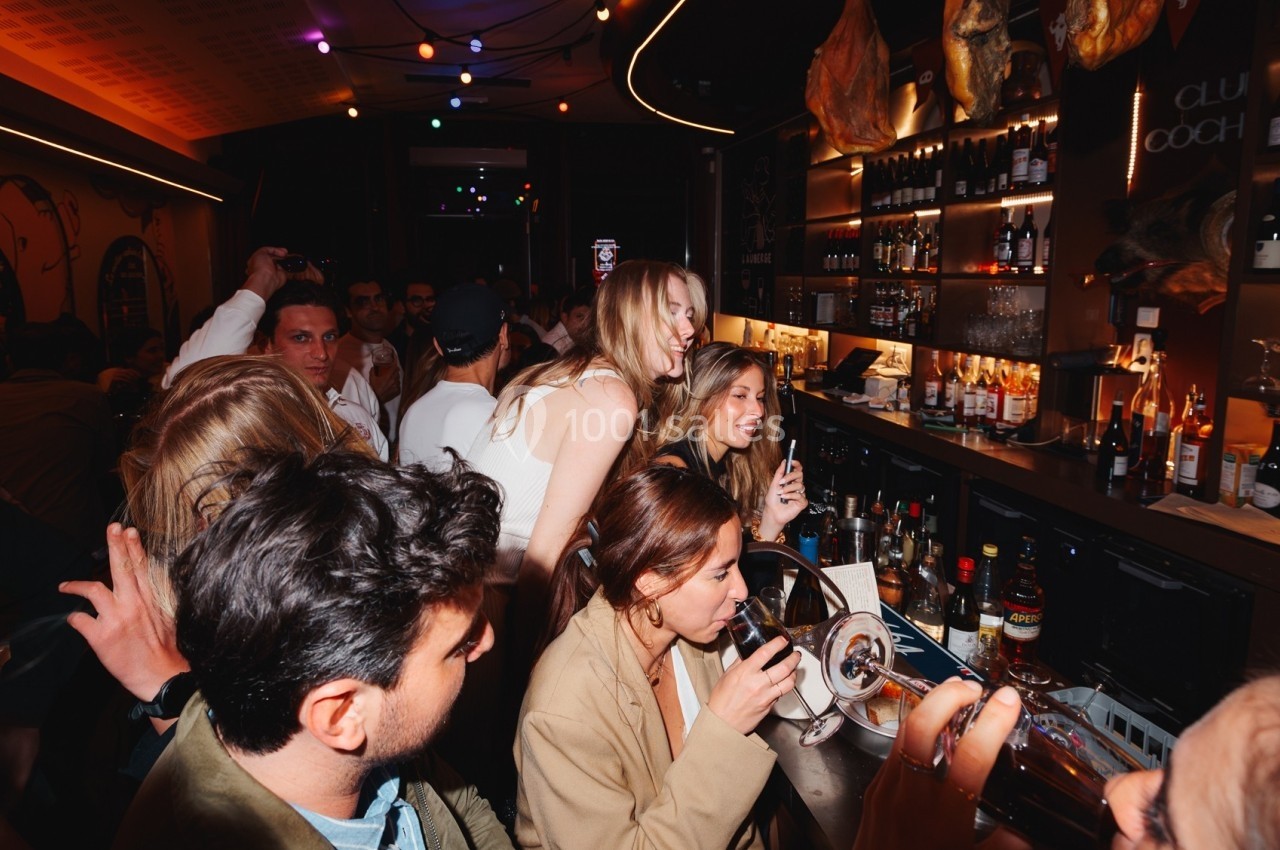 Des personnes discutent et boivent dans un bar animé, éclairé par des lumières colorées.
