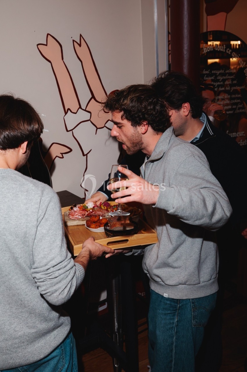 Un homme tient un verre et discute avec une personne portant un plateau de nourriture dans un lieu animé.