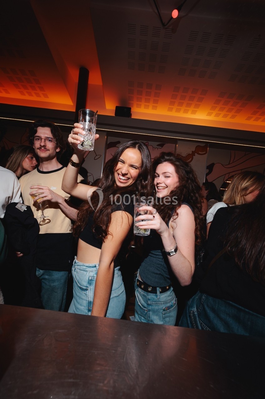 Deux jeunes femmes souriantes tenant des verres, entourées de personnes dans une ambiance festive et lumineuse.