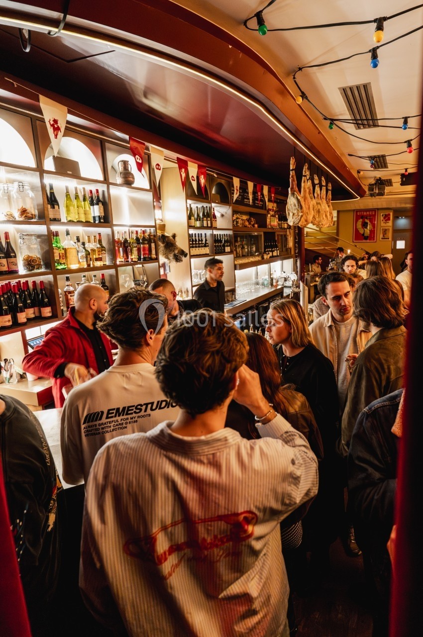 Des personnes discutent et se tiennent près d'un comptoir dans un bar animé, éclairé par des lumières chaleureuses.