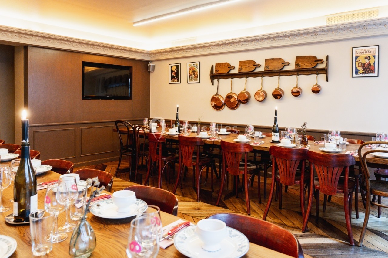 Salle de restaurant chaleureuse avec tables en bois dressées, chaises assorties et décoration murale de casseroles en cuivre.
