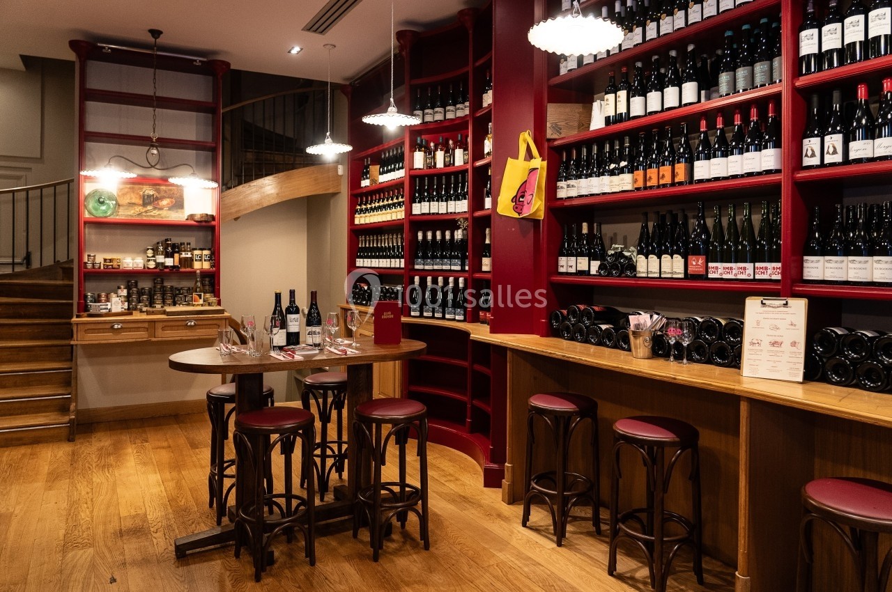 Salle d'une cave à vin avec étagères remplies de bouteilles, table haute et tabourets en bois.