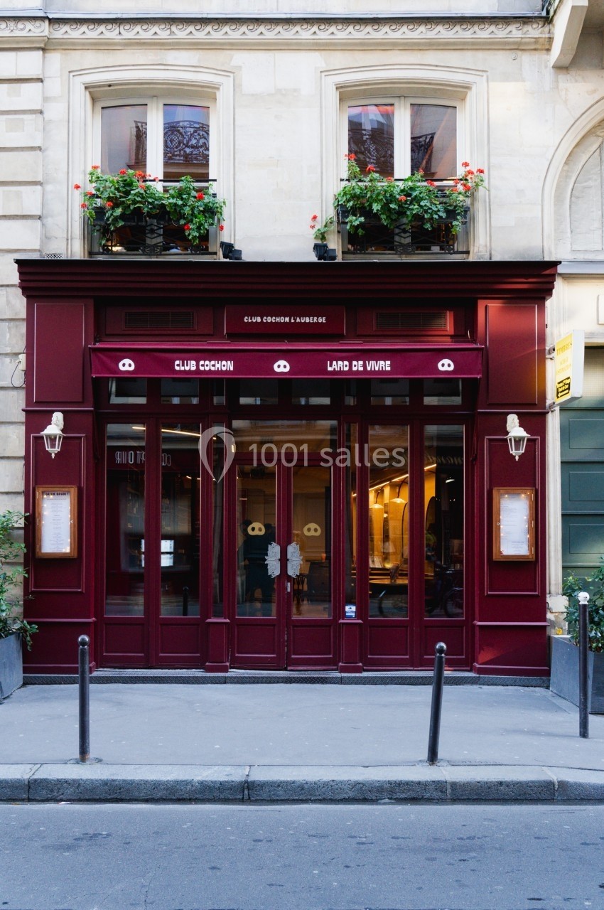Façade d'un restaurant avec une devanture rouge, deux fenêtres ornées de fleurs et une terrasse discrète.