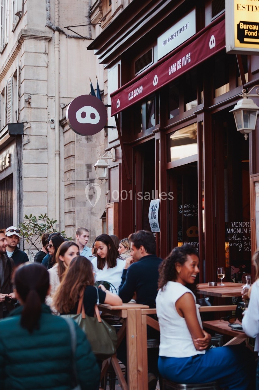 Terrasse animée d'un café en ville, avec des clients assis et discutant devant une façade en bois.