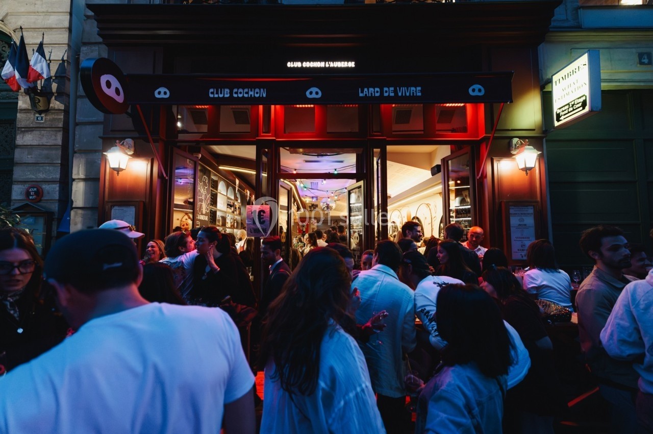Foule rassemblée devant l'entrée éclairée d'un bar-restaurant en soirée, ambiance animée et conviviale.