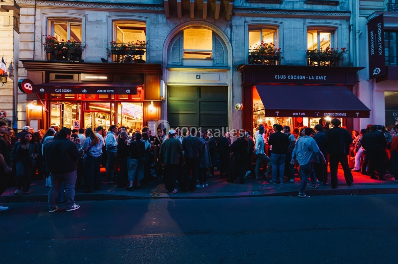 Foule rassemblée devant des bars éclairés en soirée, avec des façades parisiennes en arrière-plan.