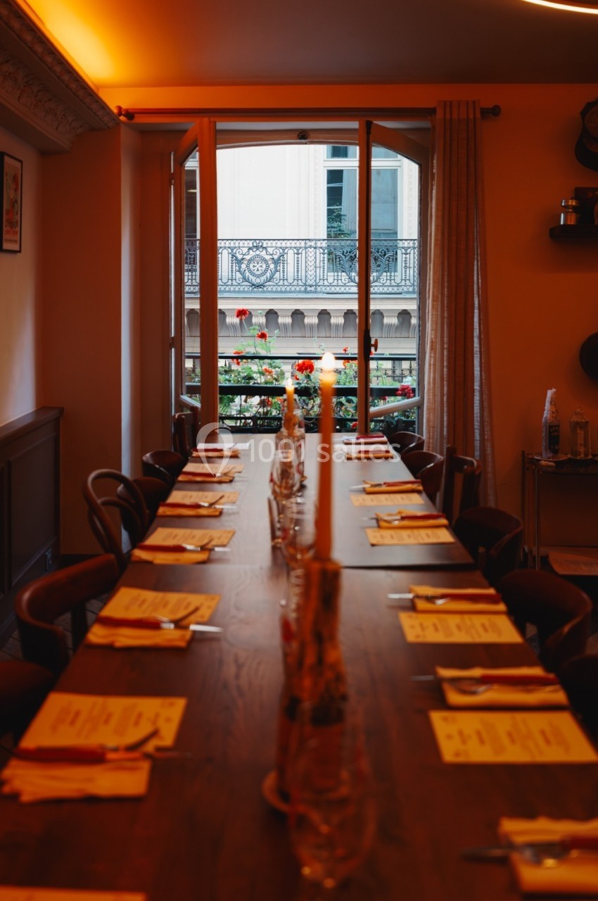 Table en bois dressée avec menus et bougies, située dans une salle chaleureuse avec vue sur un balcon fleuri.