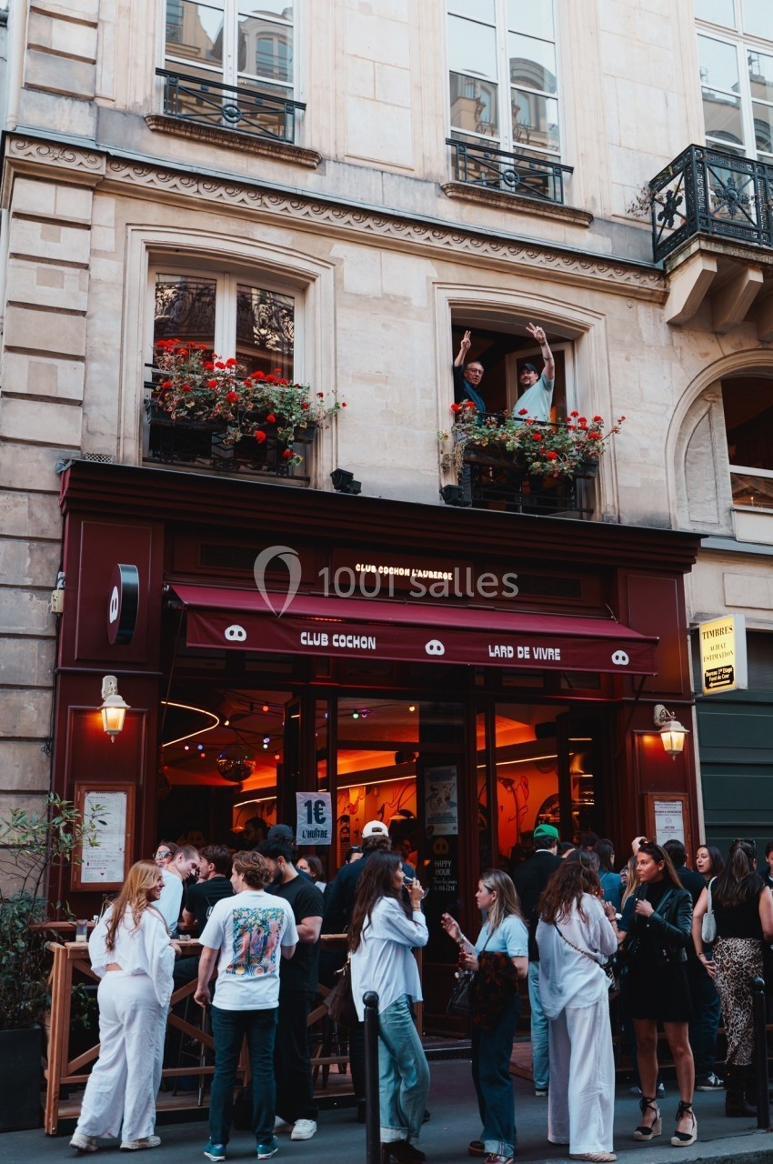 Façade d'un bar animé avec des clients en terrasse et deux personnes saluant depuis une fenêtre au-dessus.