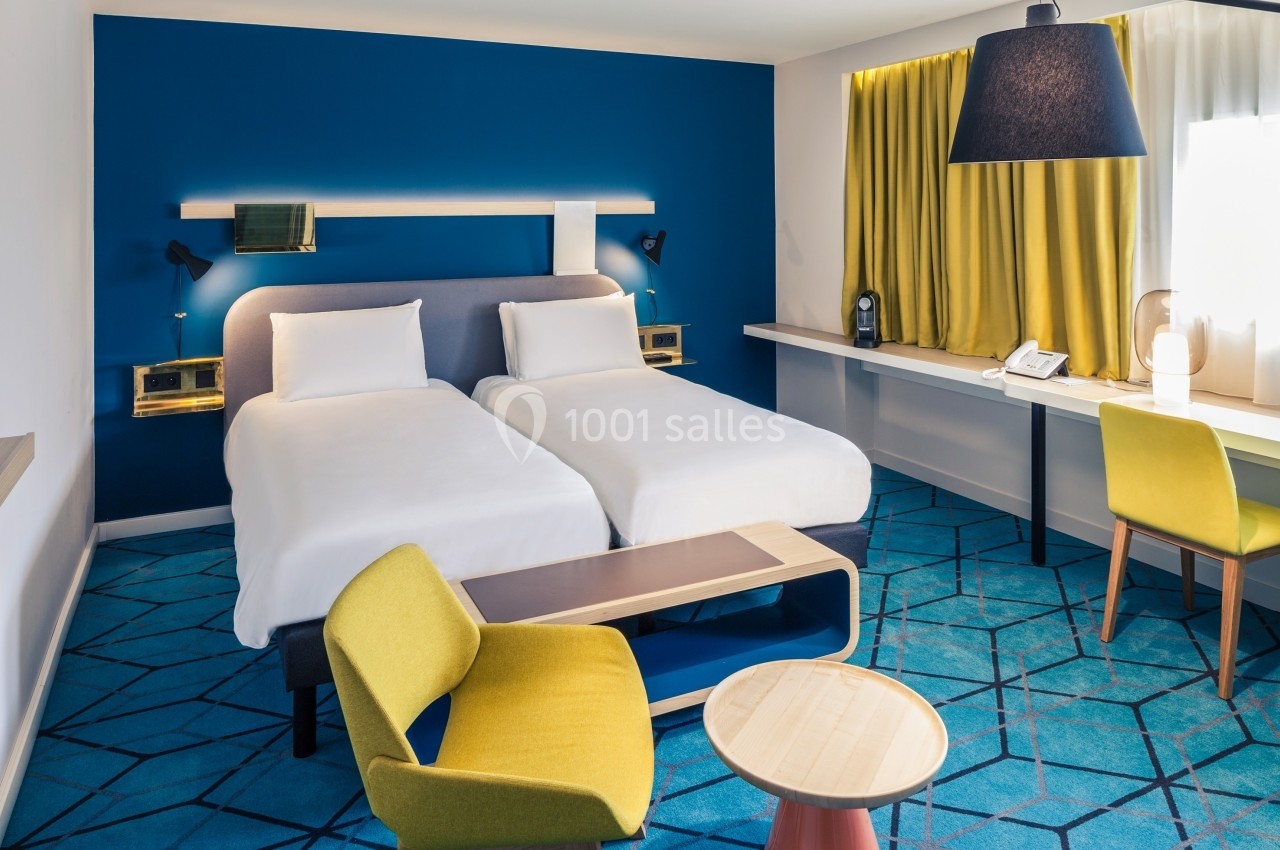 Chambre d'hôtel moderne avec deux lits simples, mobilier coloré, bureau et grande fenêtre avec rideaux jaunes.