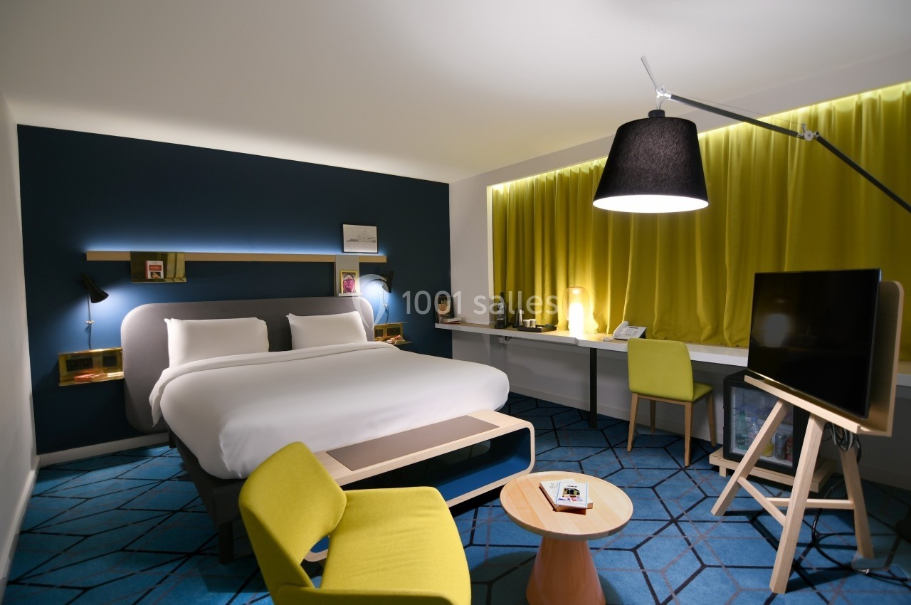 Chambre d'hôtel moderne avec lit double, bureau, fauteuils verts, éclairage tamisé et rideaux jaunes.