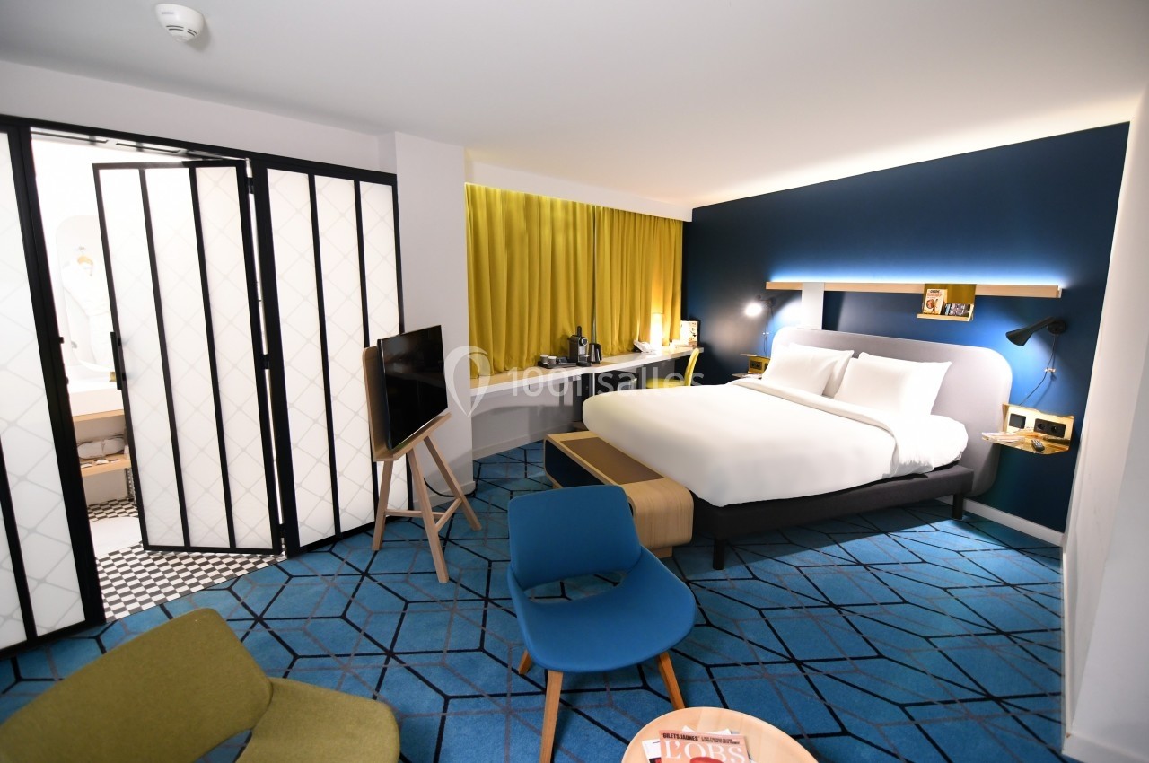 Chambre d'hôtel moderne avec lit double, bureau, télévision, fauteuils et salle de bain visible à travers une porte vitrée.