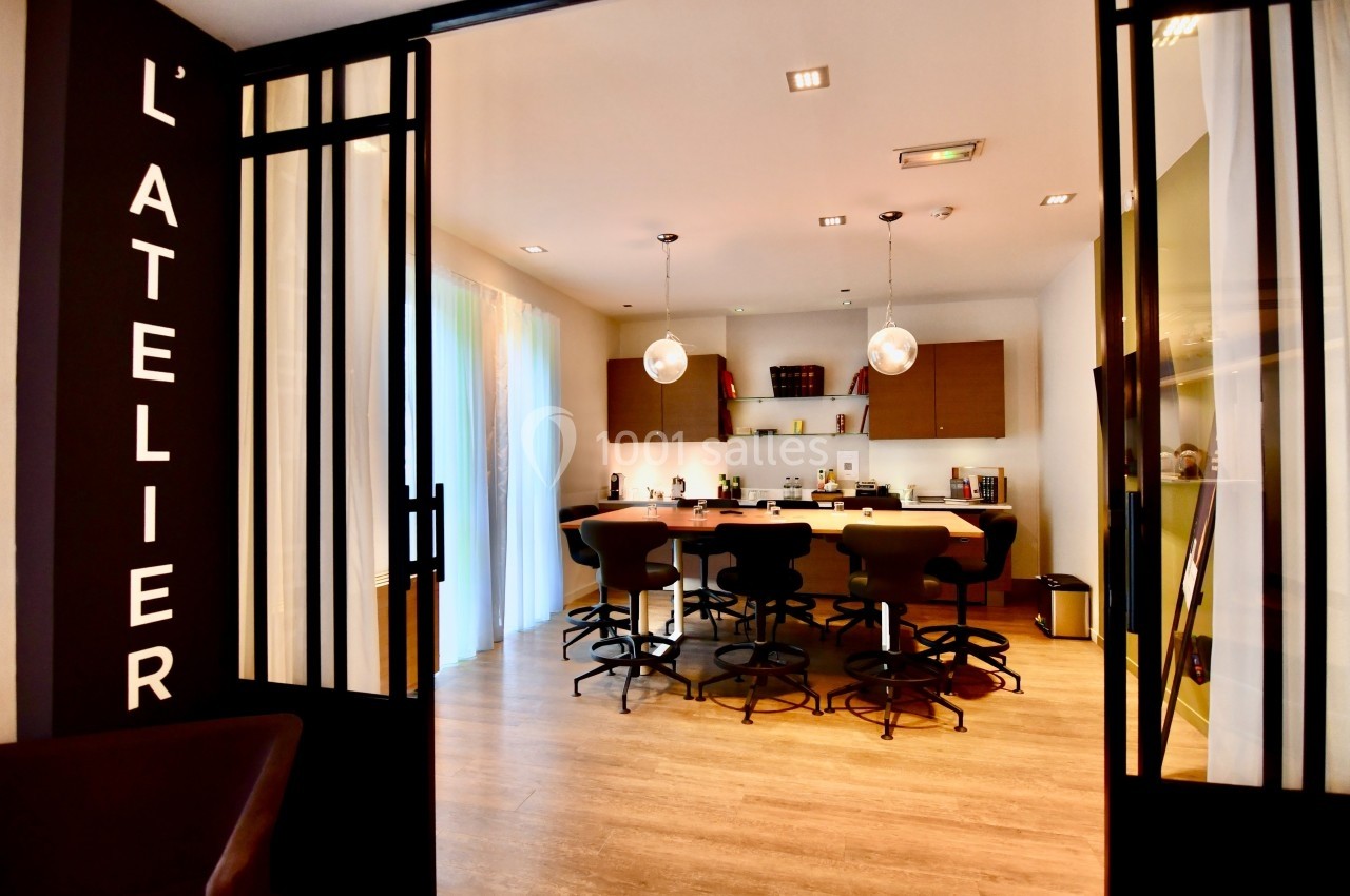 Salle de réunion moderne avec table centrale, chaises hautes et éclairage suspendu, vue depuis une entrée vitrée.