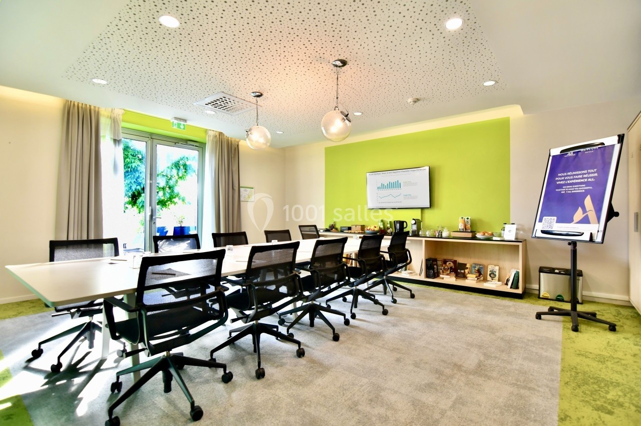 Salle de réunion lumineuse avec table rectangulaire, chaises noires, écran mural, paperboard et vue sur l'extérieur.