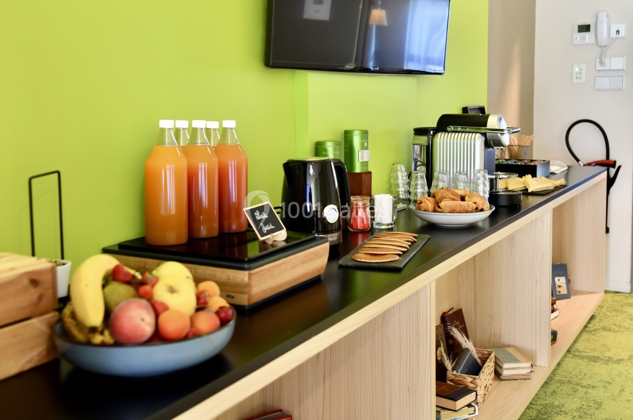 Buffet avec jus de fruits, viennoiseries, fruits frais et machine à café sur un comptoir en bois devant un mur vert.