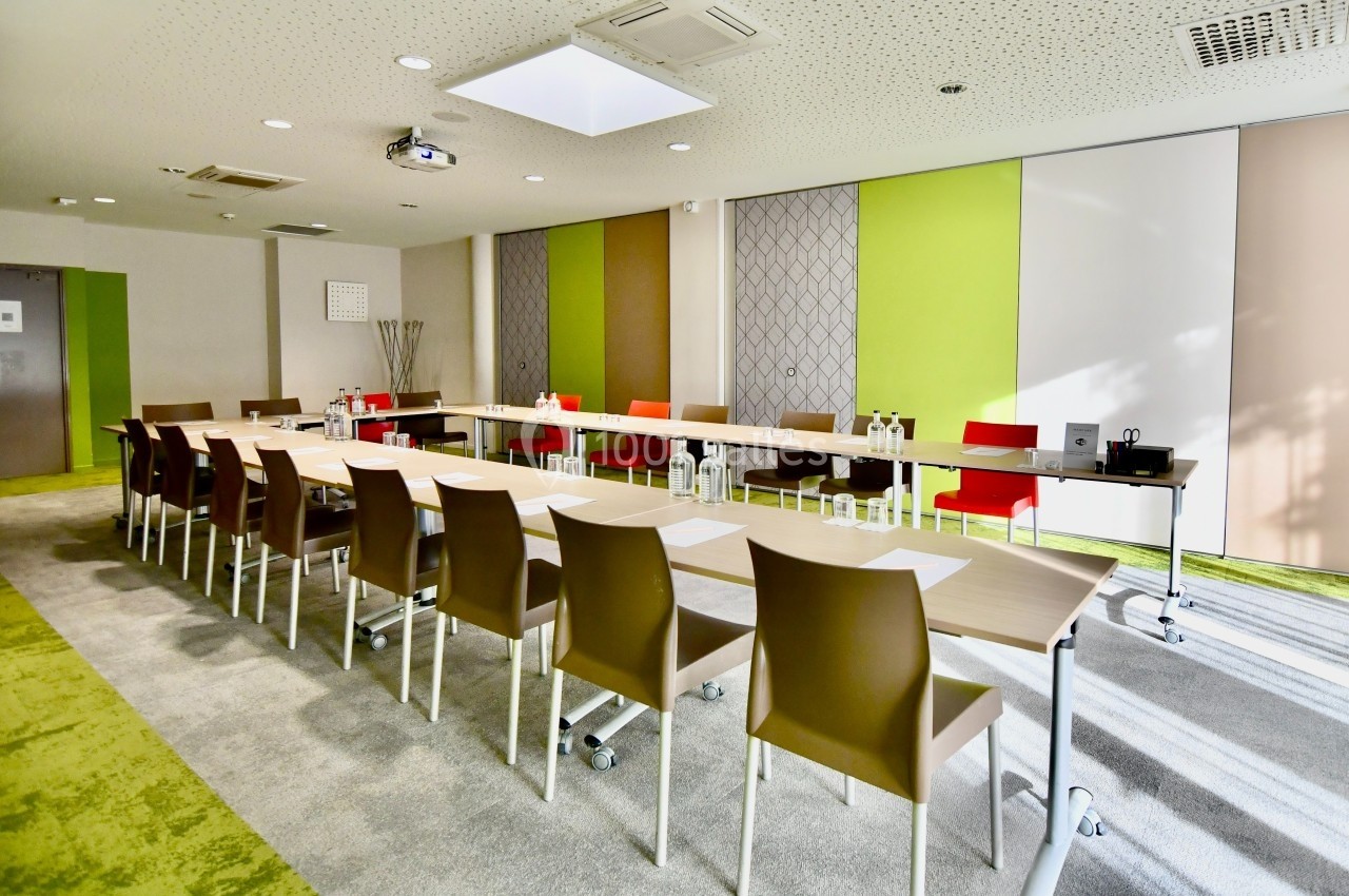 Salle de réunion lumineuse avec tables en U, chaises modernes, bouteilles d'eau et matériel de présentation.