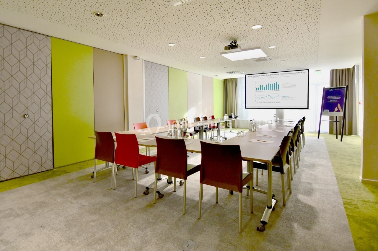 Salle de réunion lumineuse avec table rectangulaire, chaises colorées et écran affichant des graphiques.