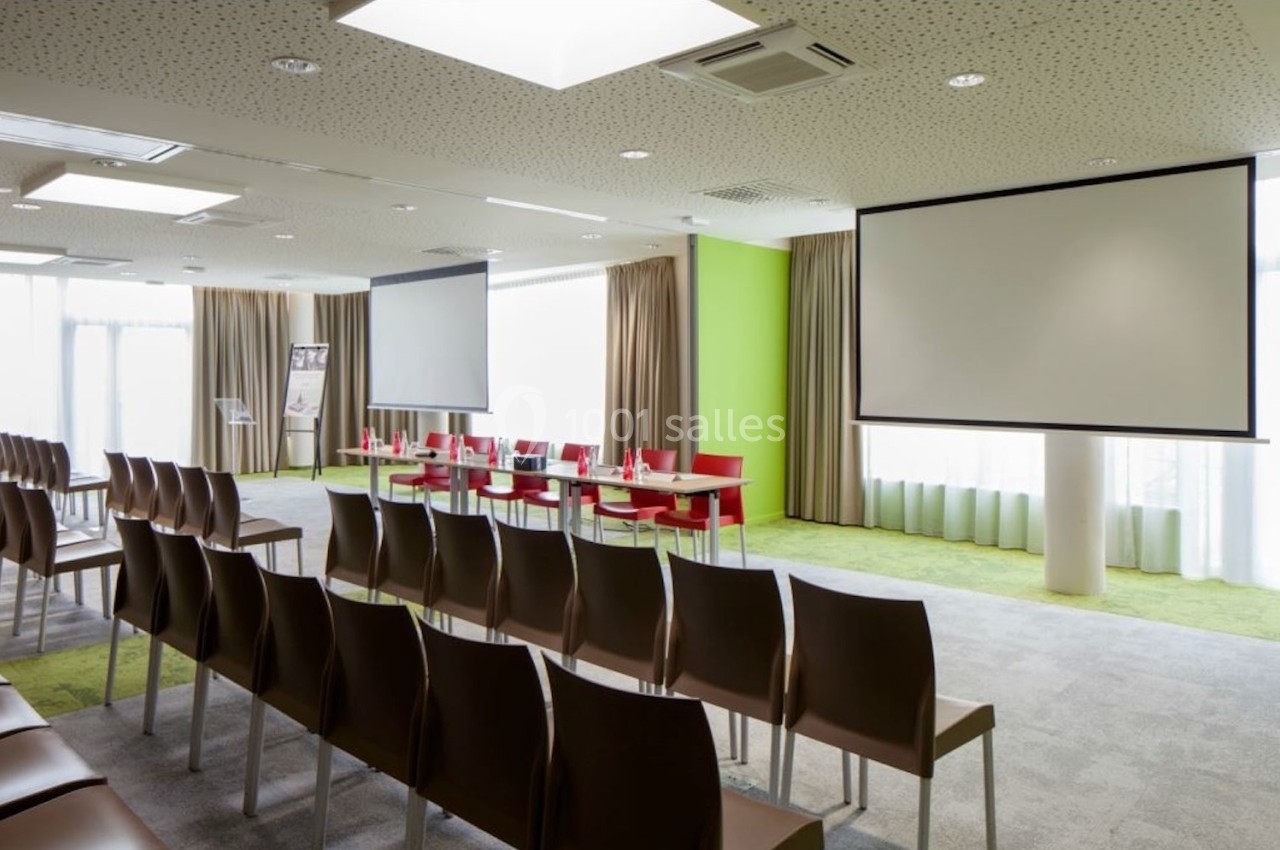 Salle de conférence lumineuse avec rangées de chaises, écran de projection et chaises rouges alignées à l'avant.