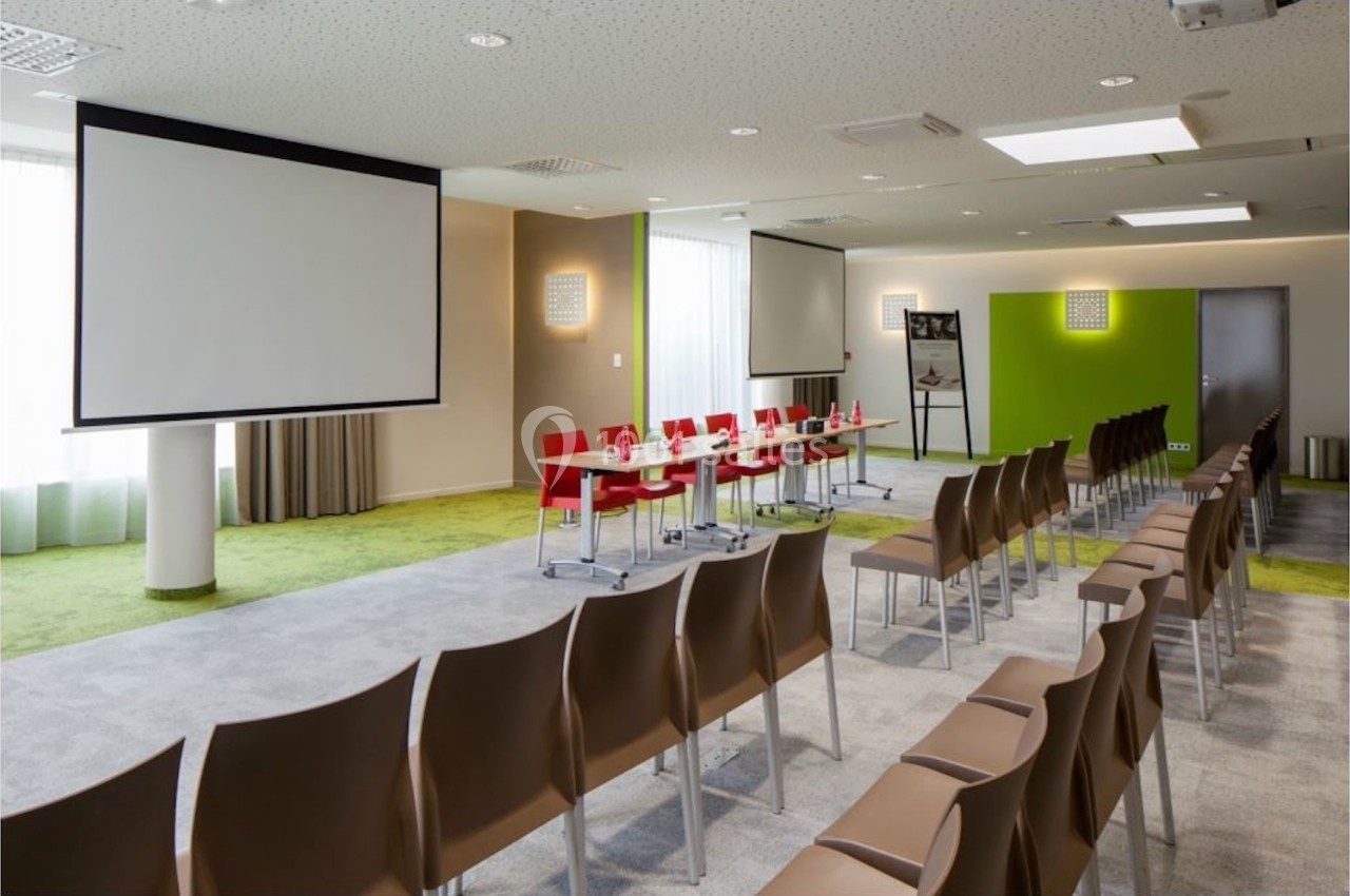 Salle de conférence lumineuse avec chaises alignées, table équipée de micros et écrans de projection.