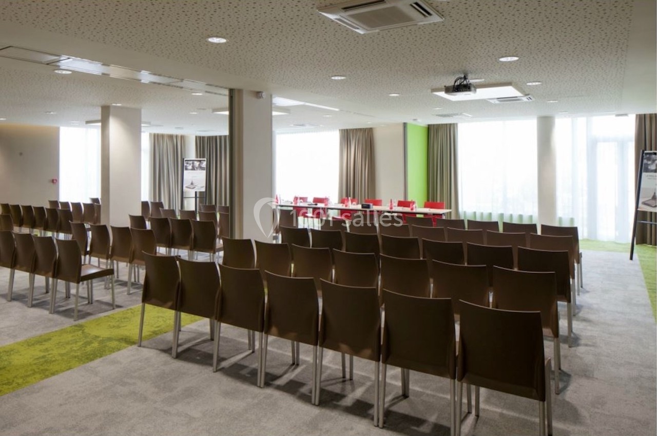 Salle de conférence lumineuse avec rangées de chaises brunes, une table et un projecteur au plafond.