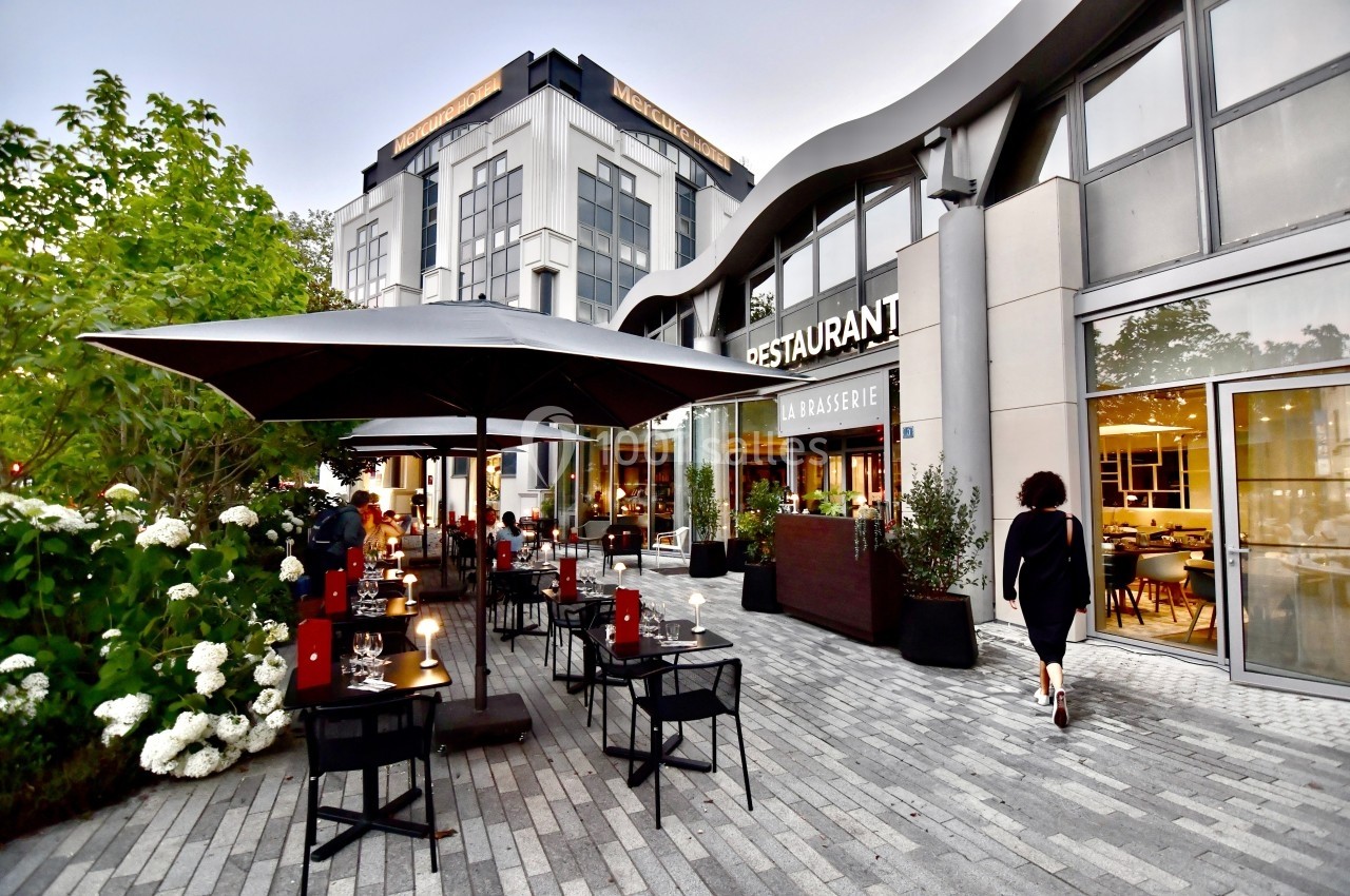 Terrasse d'un restaurant avec tables dressées, parasols et végétation, située devant un bâtiment moderne.