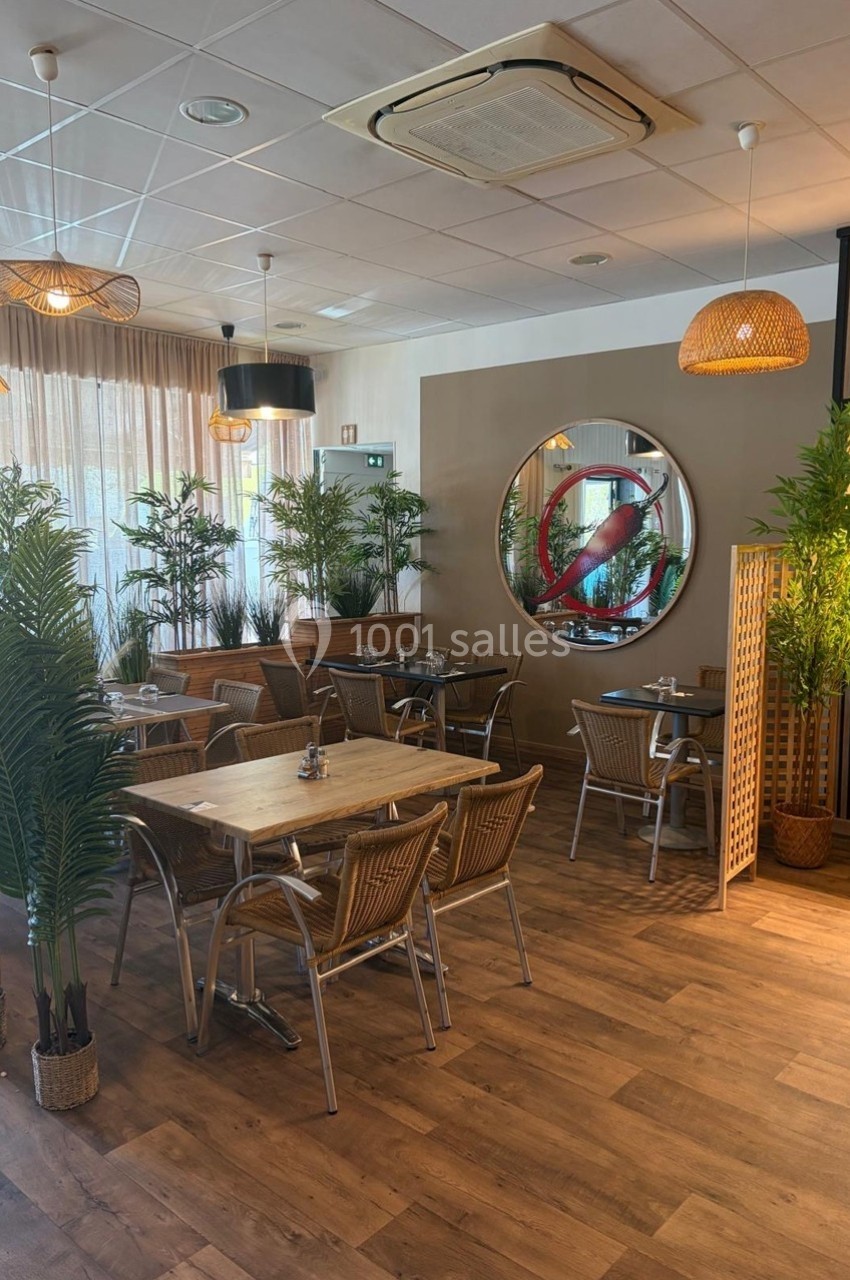 Salle de restaurant lumineuse avec tables en bois, chaises en osier, plantes décoratives et miroir mural rond.