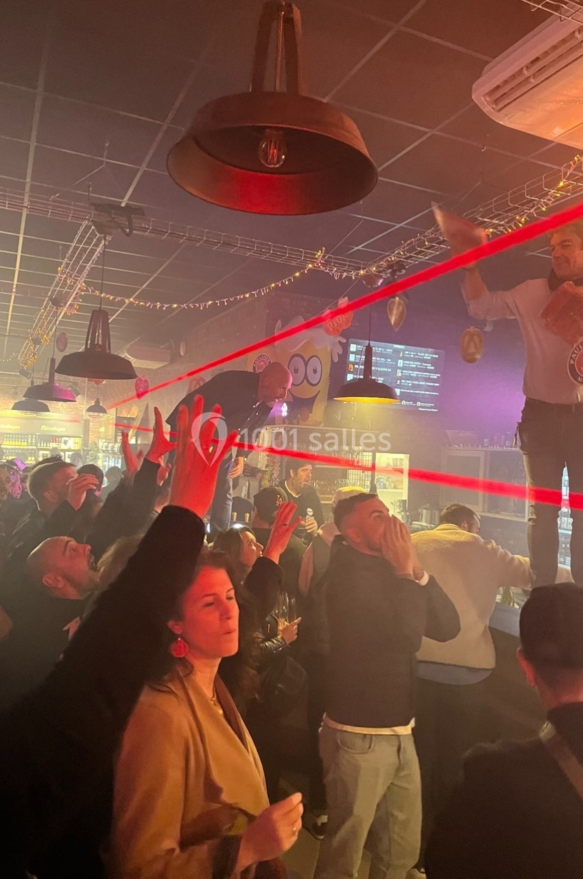 Des personnes dans un bar animé, certaines levant les bras, avec un homme debout sur le comptoir sous des lumières rouges.