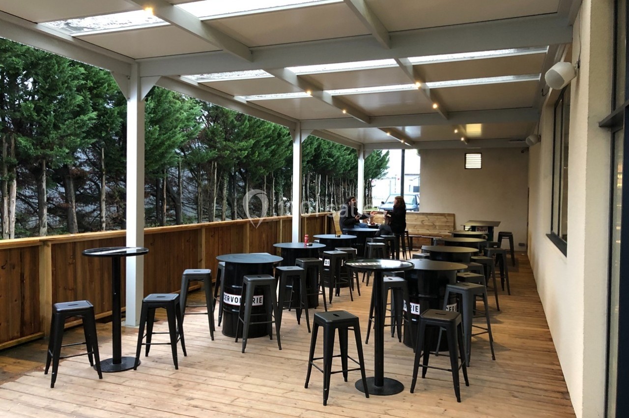 Terrasse en bois couverte avec tables hautes, tabourets noirs et deux personnes discutant au fond.