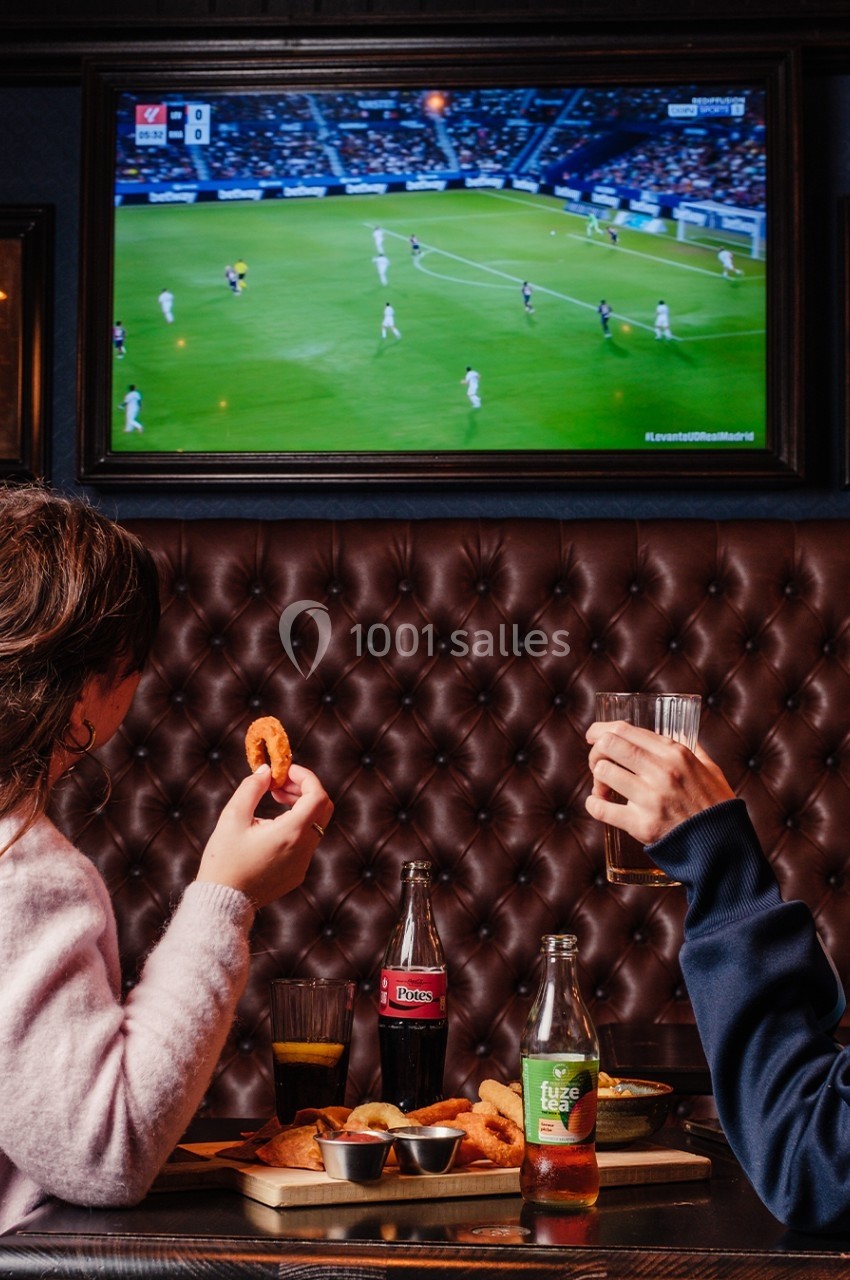 Deux personnes regardent un match de football à la télévision dans un bar, en mangeant et buvant à une table.