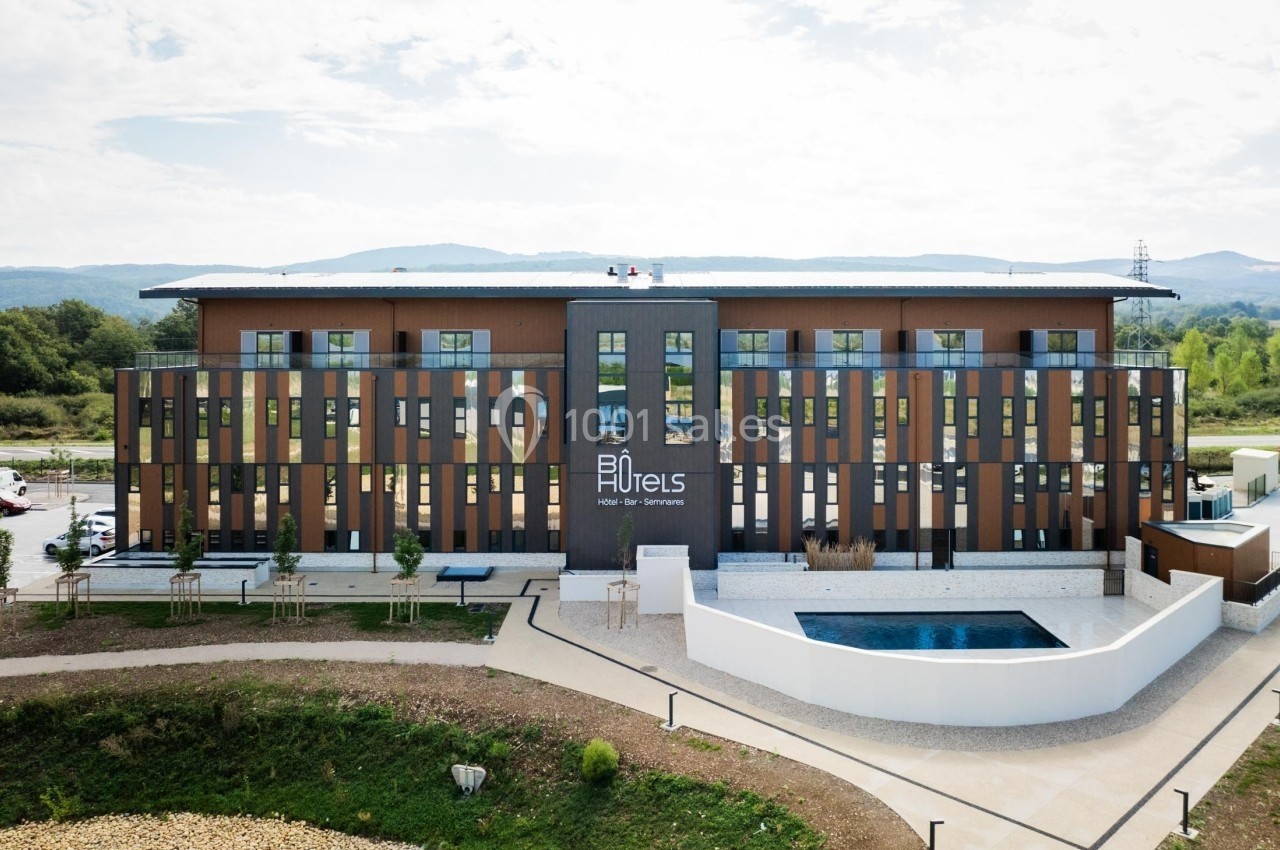 Façade d'un hôtel moderne à trois étages avec piscine extérieure, entouré de verdure et d'un ciel partiellement nuageux.