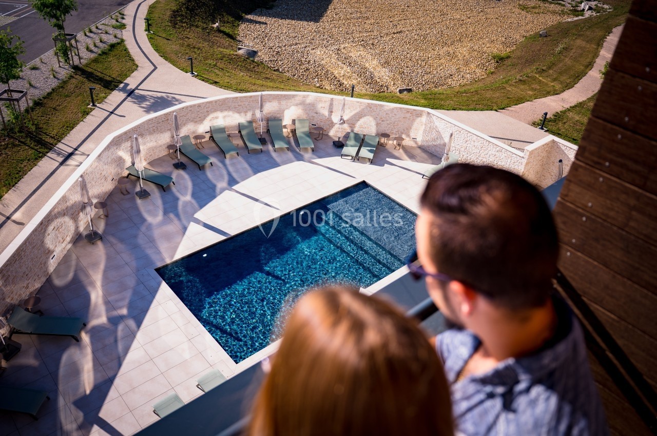 Vue plongeante sur une piscine extérieure entourée de transats, observée depuis un balcon par deux personnes.