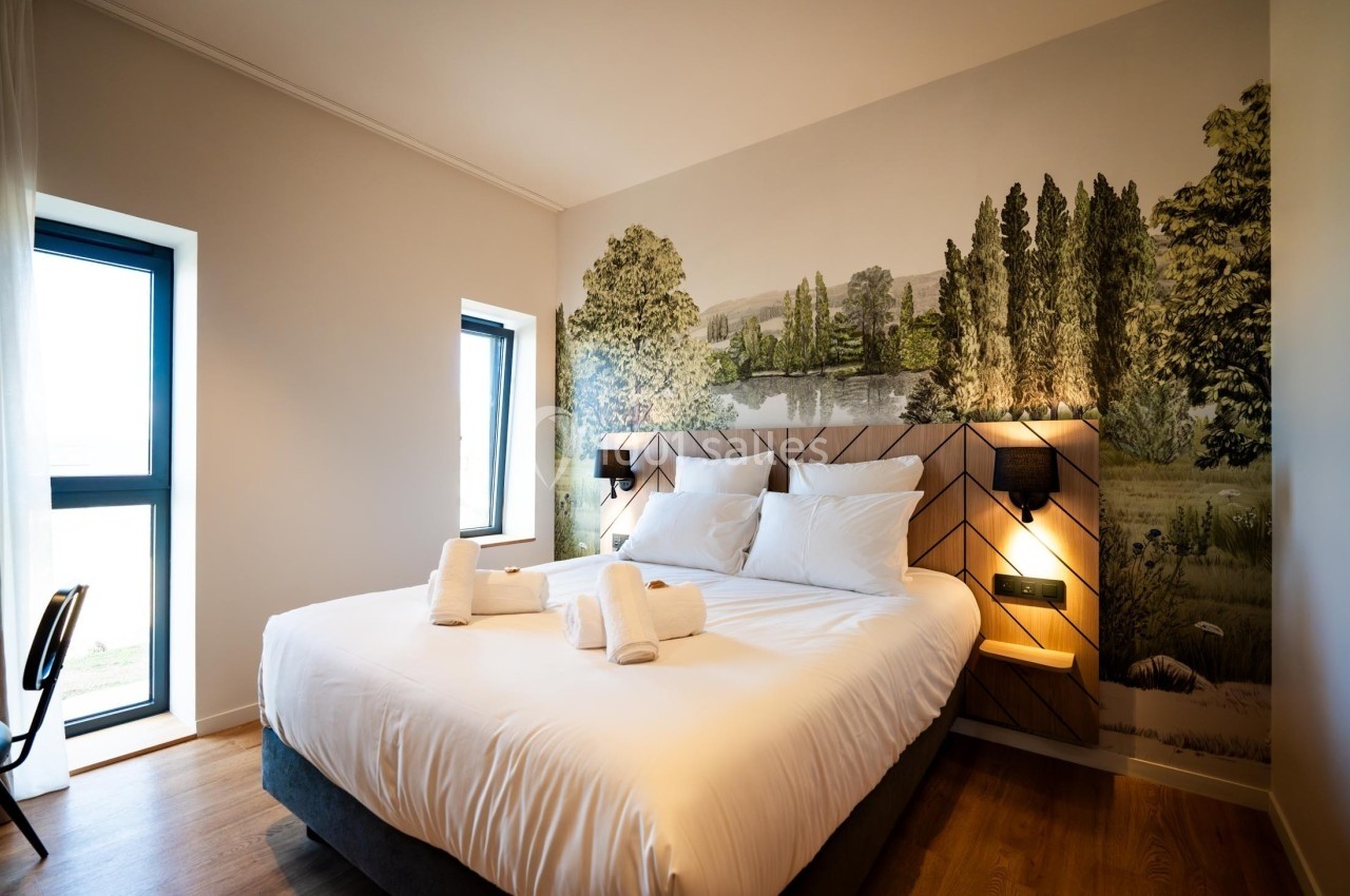 Lit double avec draps blancs, serviettes pliées, éclairage mural et fresque murale représentant un paysage naturel.