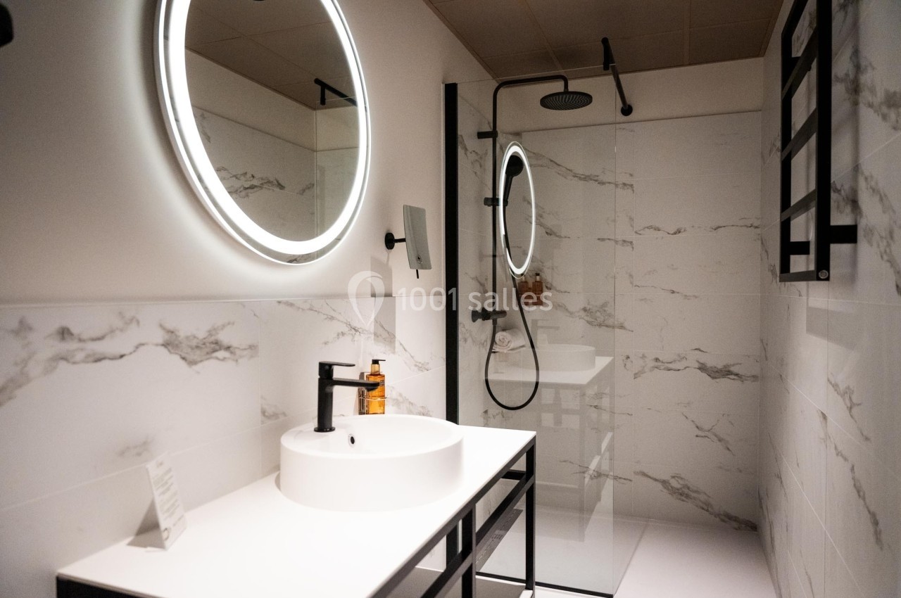 Salle de bain moderne avec lavabo blanc, miroir rond éclairé, douche à l'italienne et murs en carrelage effet marbre.
