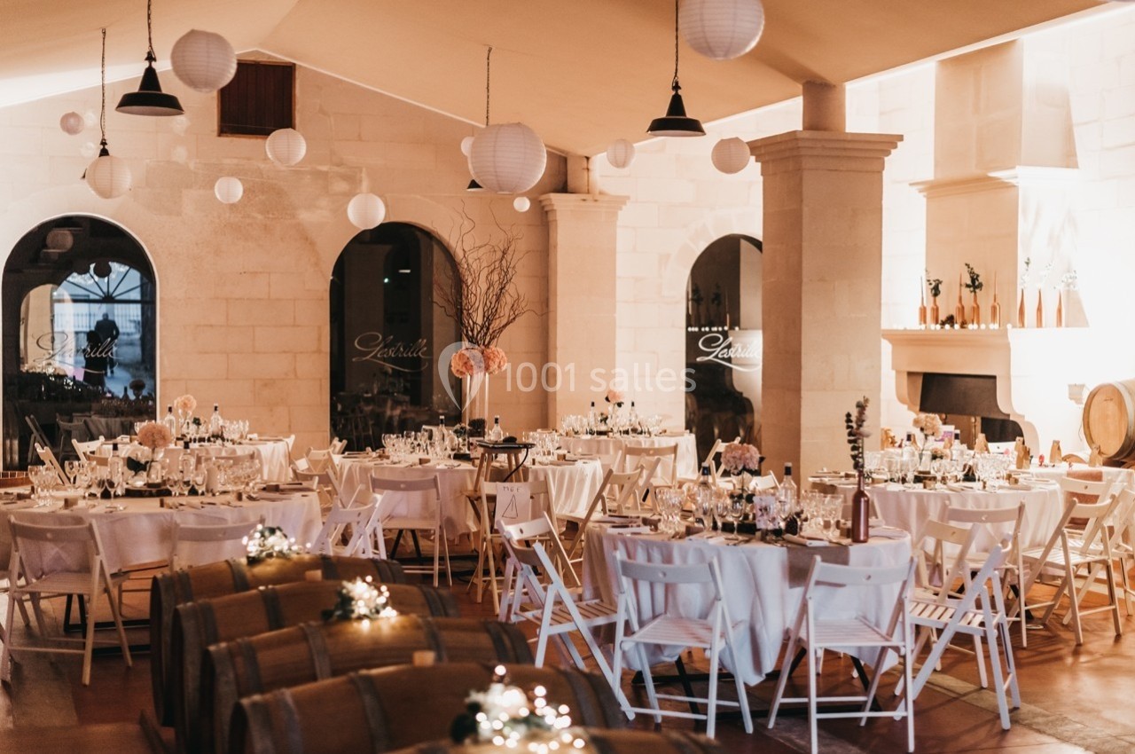 Salle de réception décorée pour un événement, avec tables dressées, chaises blanches et lanternes suspendues.