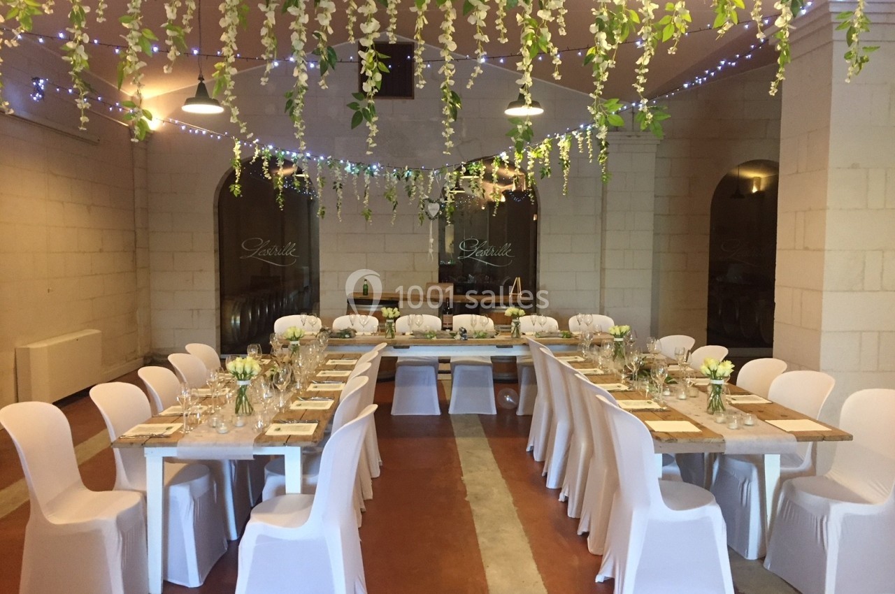 Salle décorée pour un dîner, avec tables alignées, chaises blanches et guirlandes lumineuses suspendues.