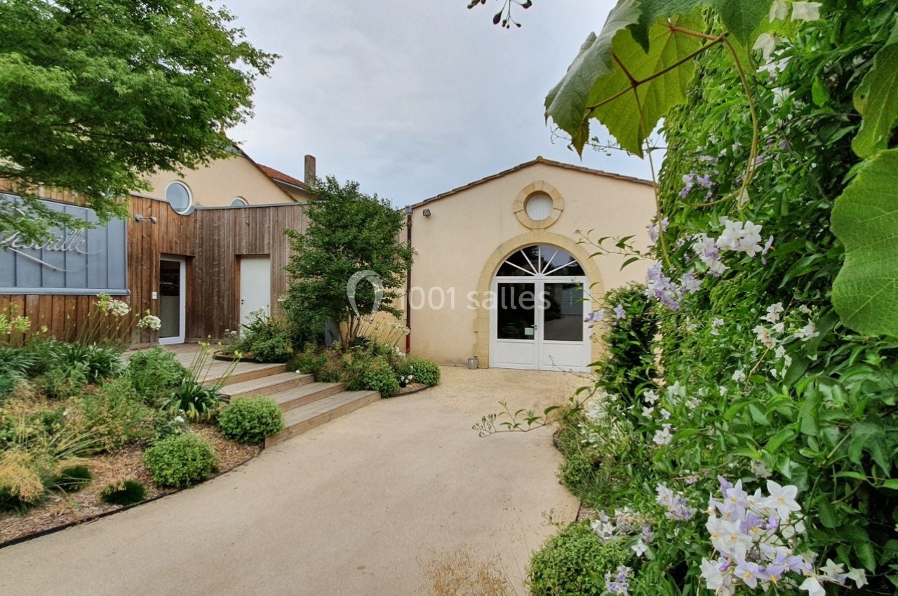 Entrée d'un bâtiment entourée de végétation, avec un chemin en béton et des plantes grimpantes sur la droite.