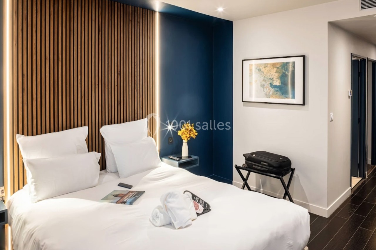 Chambre d'hôtel moderne avec lit double, mur bleu, décoration minimaliste et valise posée sur un support.