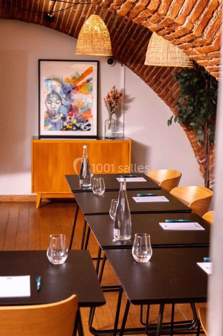 Salle de réunion avec tables noires, chaises en bois, bouteilles d'eau, carnets, et une décoration murale colorée.